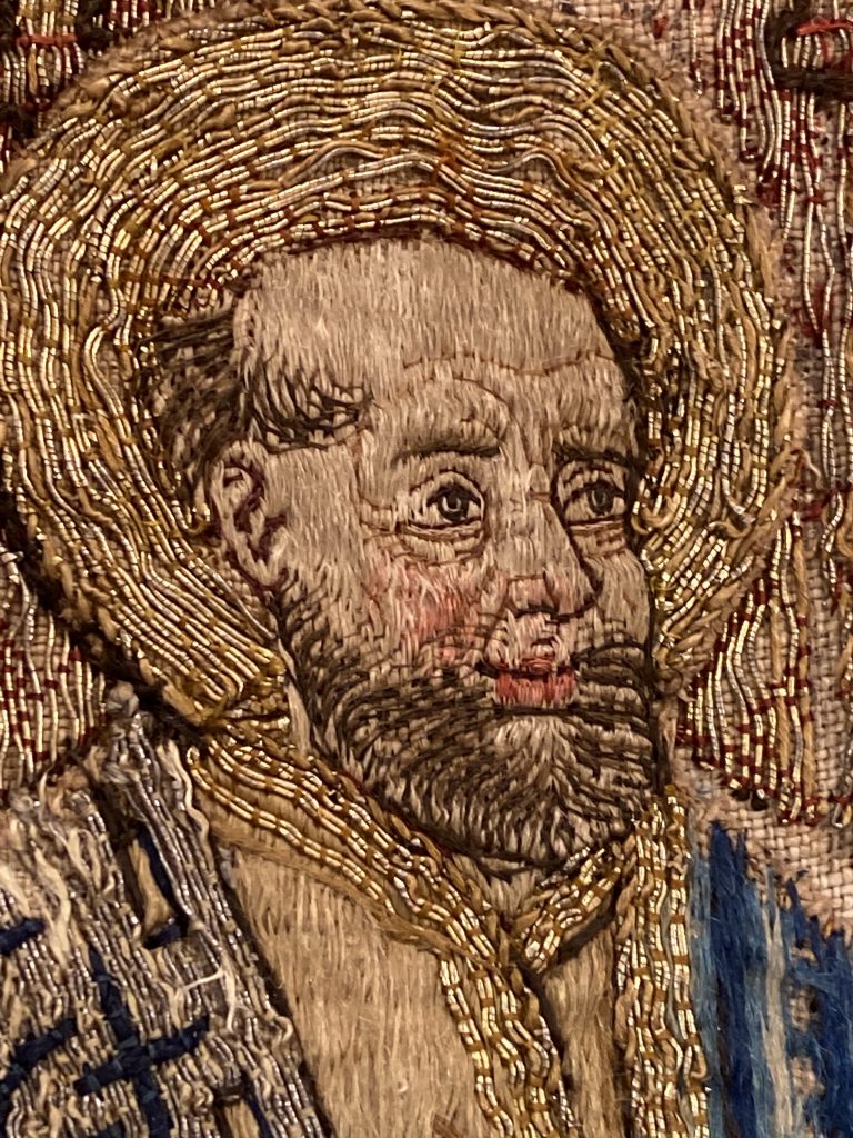 En närbild på ett broderat ansikte. Mannen har skägg, en tonsur och en gloria runt huvudet.