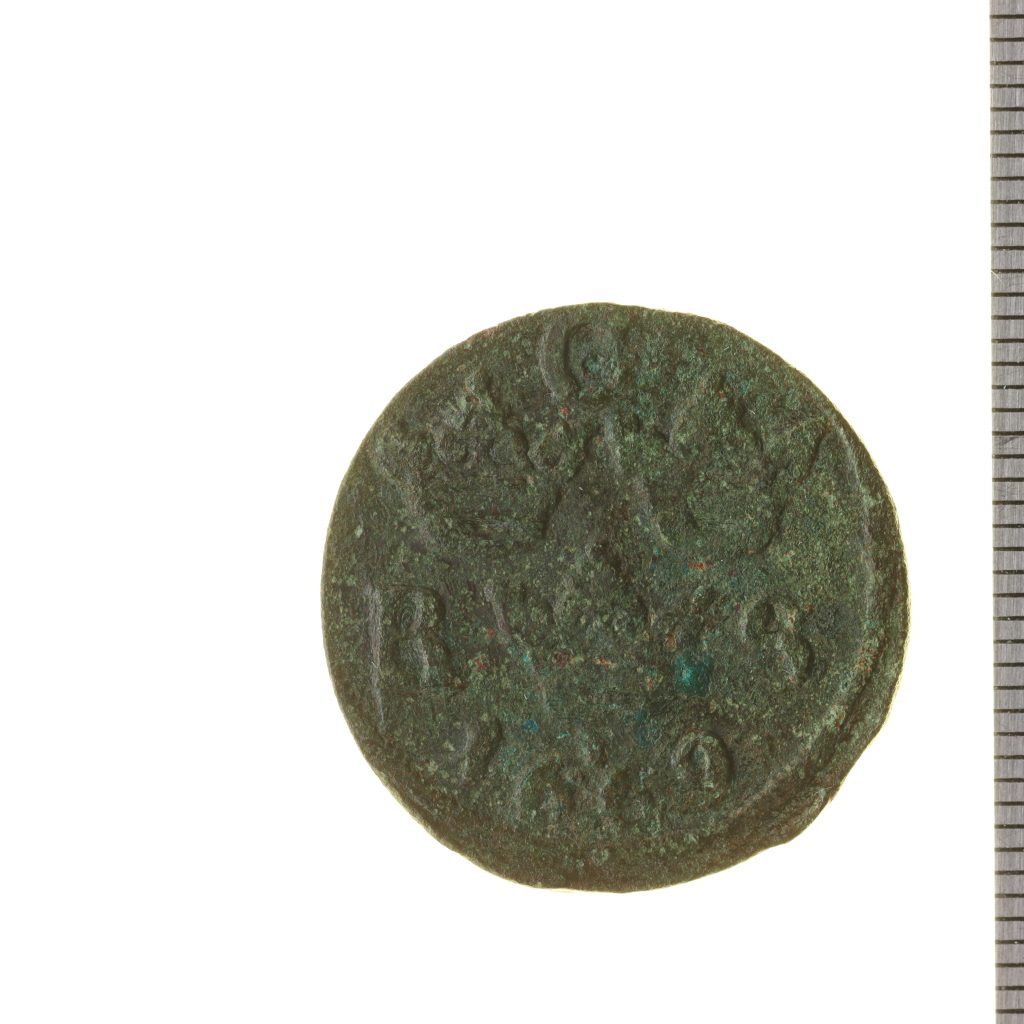 Bilden visar ett gammalt kopparmynt med en mätsticka vid sidan av myntet. 