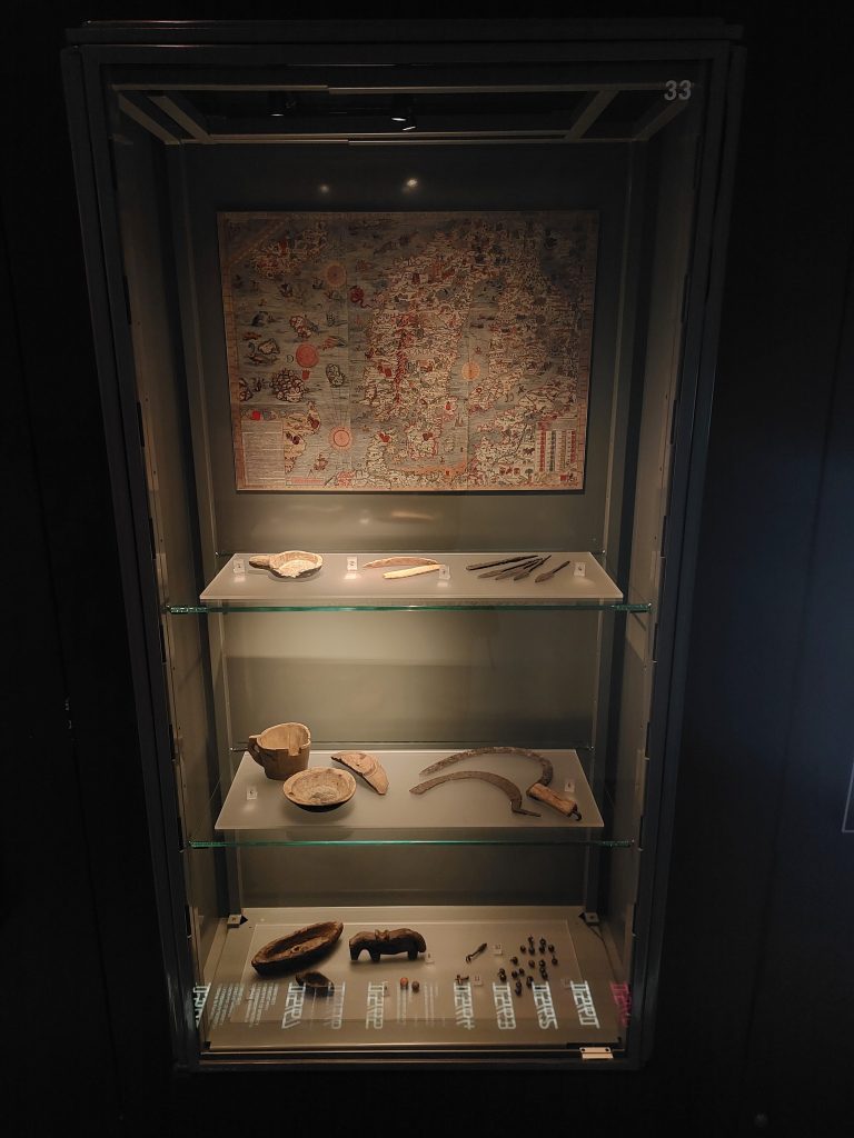 Bild på monter som visar senmedeltida föremål som berättar om människans vardagsliv, bland annat träkärl, skäror, pilspetsar, knappar och leksaker tillverkade av bark.