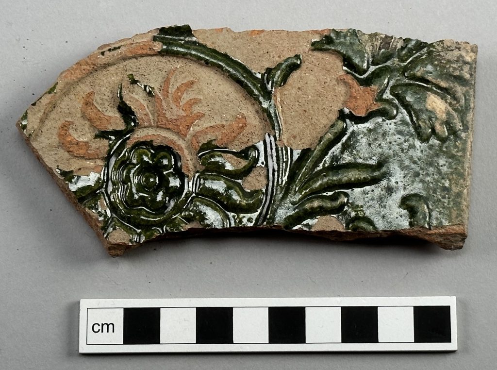 Bilden visar en fragmenterad kakelplatta. Kakelplattan är dekorerad med blommotiv, kanske en nejlika, och är täckt med grön glasyr. Glasyren är avskavd på delar av kaklet.