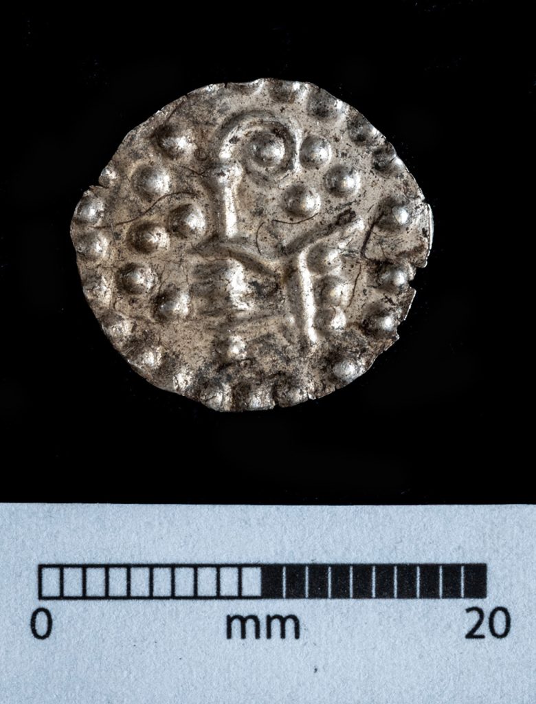 Silvermynt med en hand som håller i en stav som är ihoprullad i toppen. Myntets yta täcks av konvecka prickar.