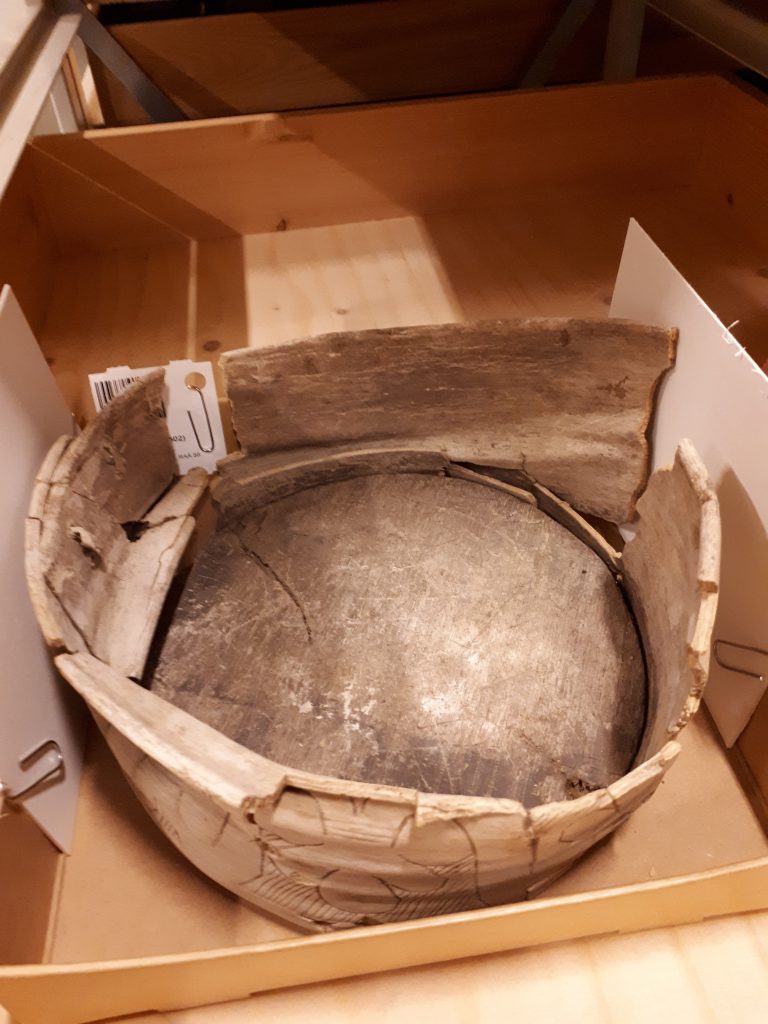 Svepask med ihopsjunkna sidor och med ena sidan lossad, placerad i en brun ask i en föremålsback av trä.