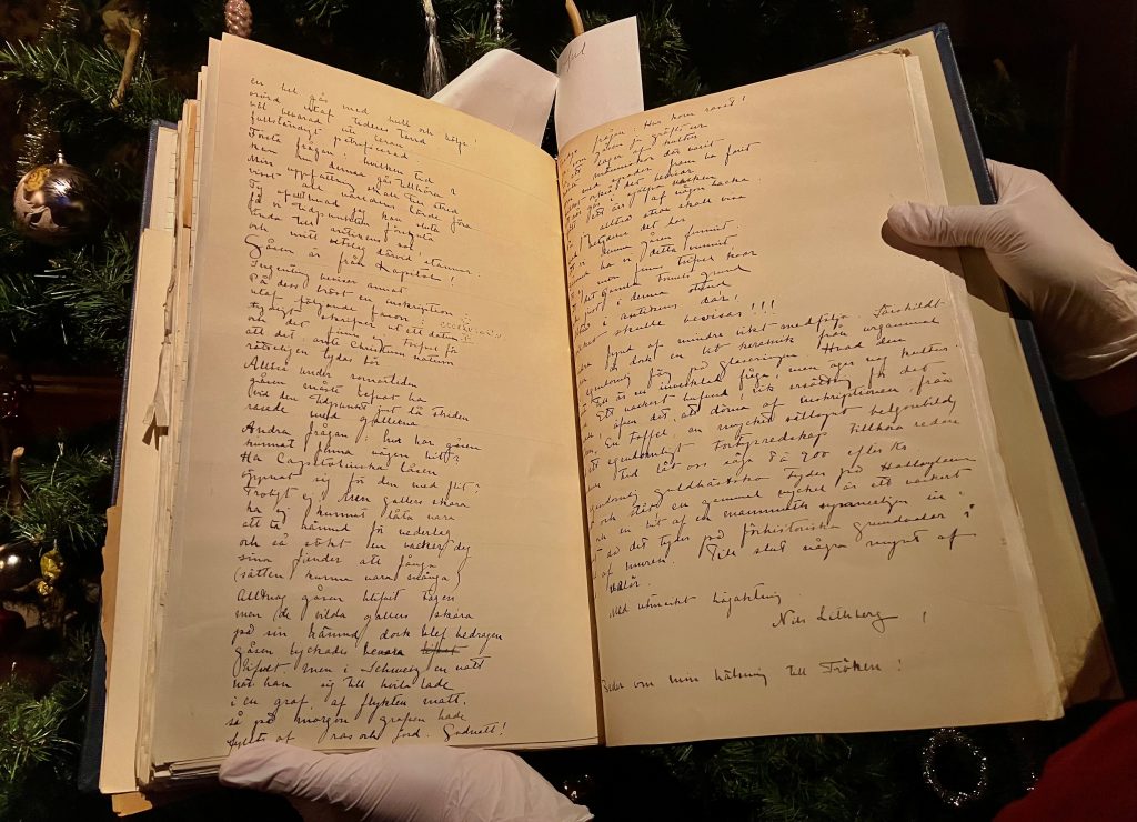 En stor bok med handskriven text hålls uppslagen av två händer med vita handskar.