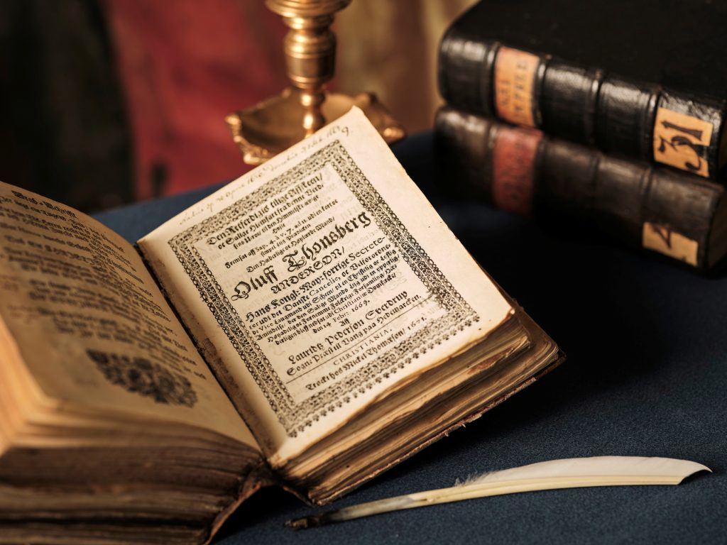 En gammal bok ligger uppslagen på ett bord. I bakgrunden ser man fler böcker och en ljusstake.