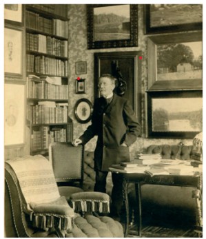 Svartvitt foto av en man som står i ett rum med bokhyllor och tavlor på väggarna.