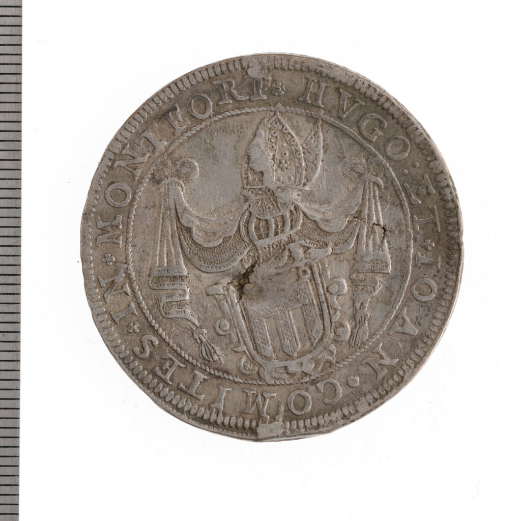 Silverfärgat mynt med bild av en vapensköld och texten "Hugo et Ioan comites in Montfort"